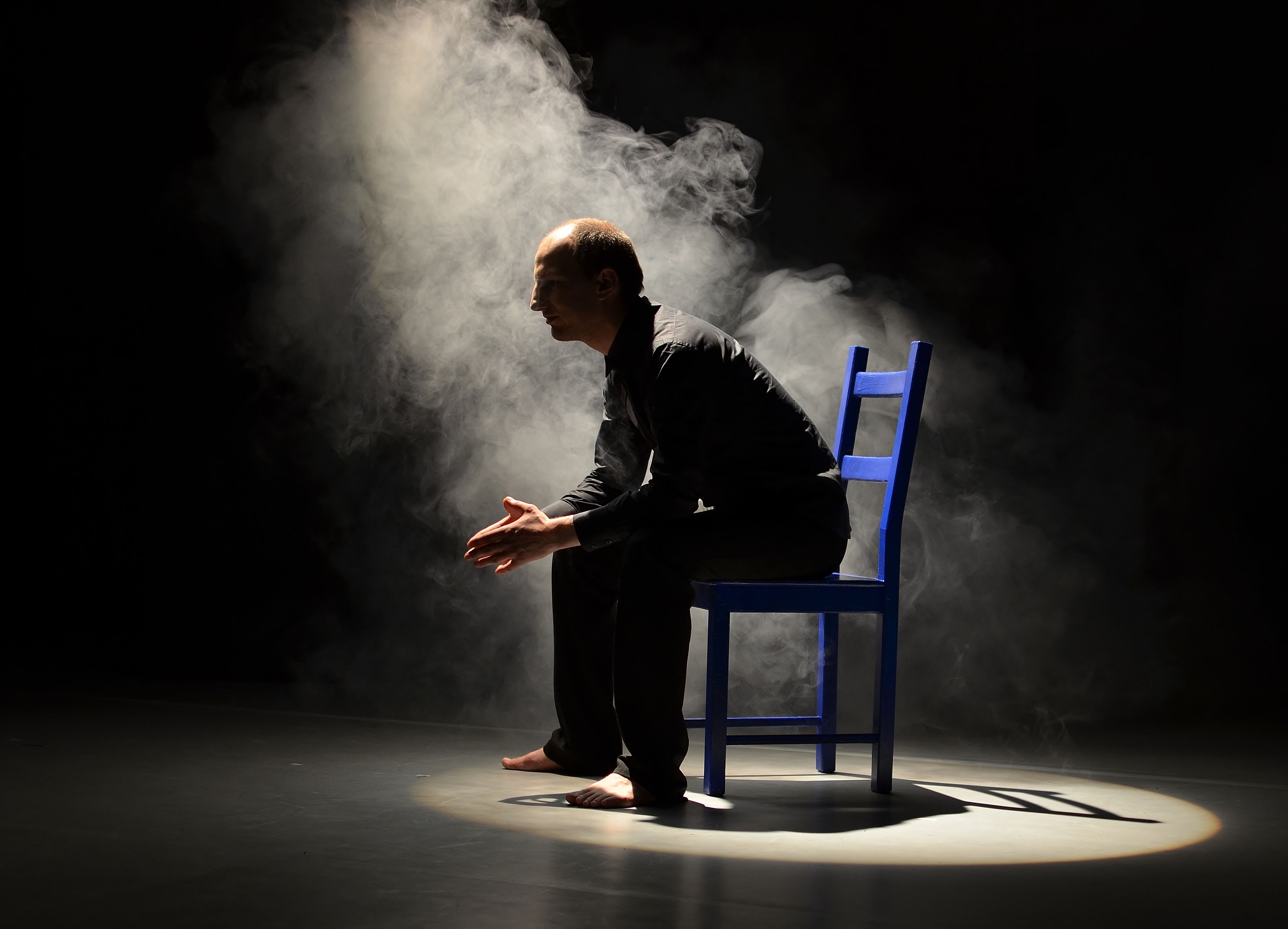 Dunkle Bühne, ein einziger Lichtstrahl fällt auf einen auf einem blauen Stuhl sitzenden Mann, der barfuß und schwarz bekleidet ist